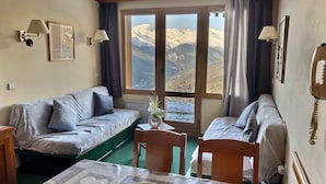 A cozy room with a sofa, a dining table, and a view of mountains through large windows. - APARTMENT AT THE FOOT OF PISTE 4/5 PEOPLE (LA PLAGNE-TARANTAISE)