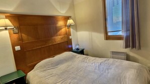 A bedroom with a wooden headboard, a bed with a white duvet, a nightstand with a lamp, and a window with blinds. - APARTMENT AT THE FOOT OF PISTE 4/5 PEOPLE (LA PLAGNE-TARANTAISE)