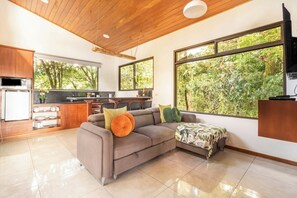 A modern kitchen with a built-in oven, a dining area with a sofa and a coffee table, and large windows offering a view of greenery.