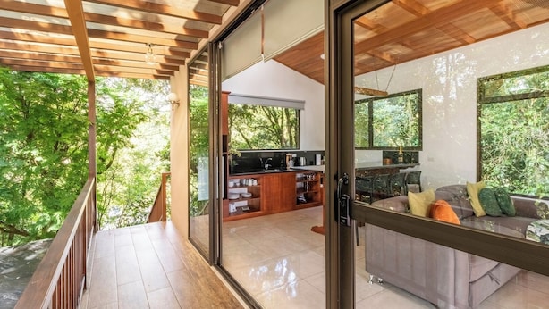 A modern kitchen with a large island, a dining area with a sofa, and a sliding glass door leading to a balcony with a wooden deck. - Sunset Views Over the Cloud Forest • Steps from Zip Lines, Farm & Wine (Monteverde)