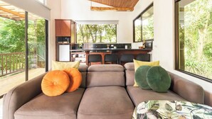 A modern living area with a sofa, cushions, and a table. A kitchen is visible in the background with a dining area. - Sunset Views Over the Cloud Forest • Steps from Zip Lines, Farm & Wine (Monteverde)