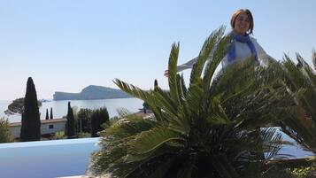 Une personne se tient près d’une piscine, entourée de palmiers, avec une vue sur une étendue d’eau et un paysage lointain.