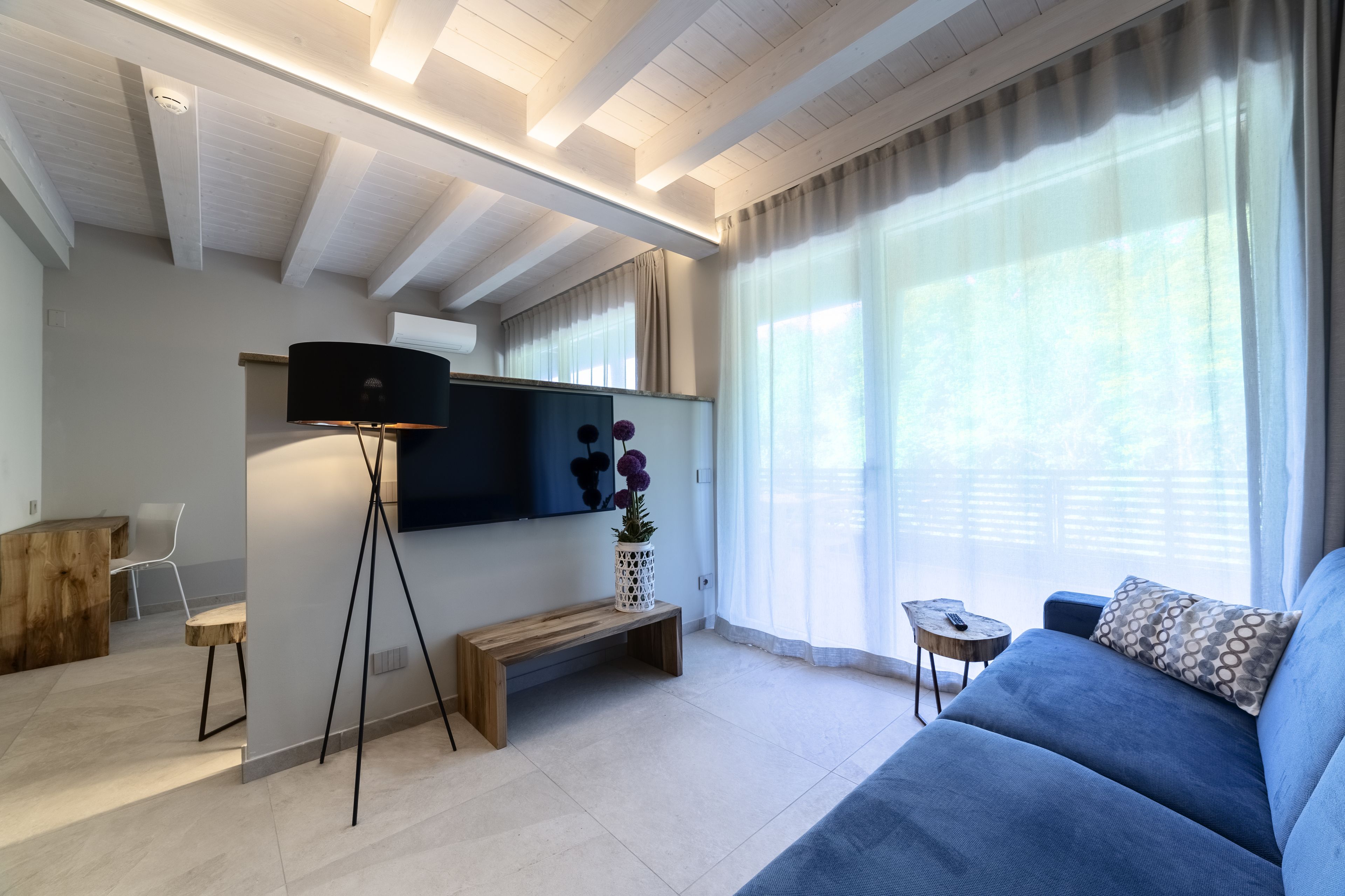 Un salon moderne avec un grand téléviseur à écran plat, un canapé bleu, un banc en bois et une petite table d’appoint.