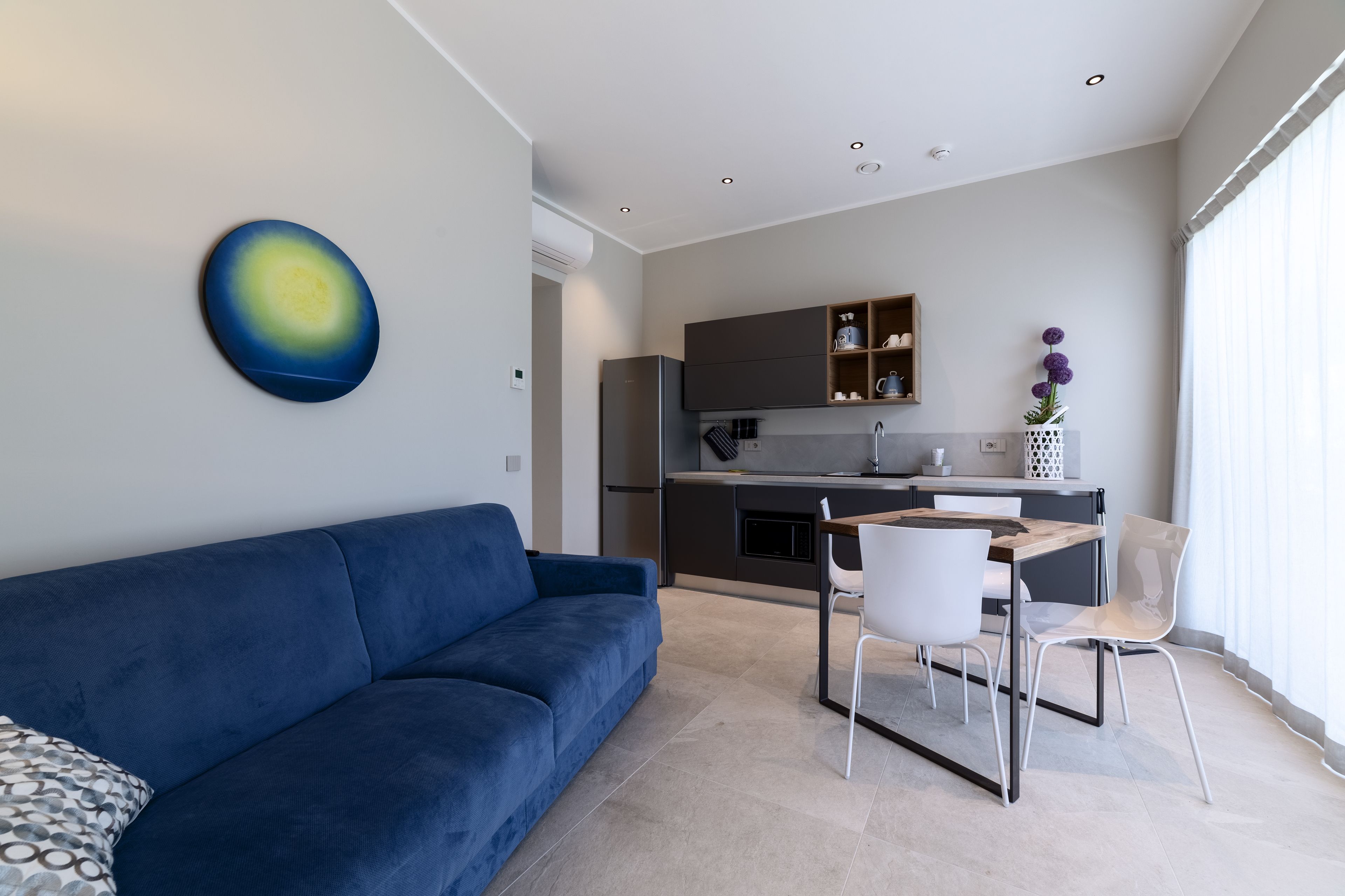 A modern living room with a blue sofa, a dining area with a wooden table and white chairs, and a kitchen with dark cabinets and a refrigerator.