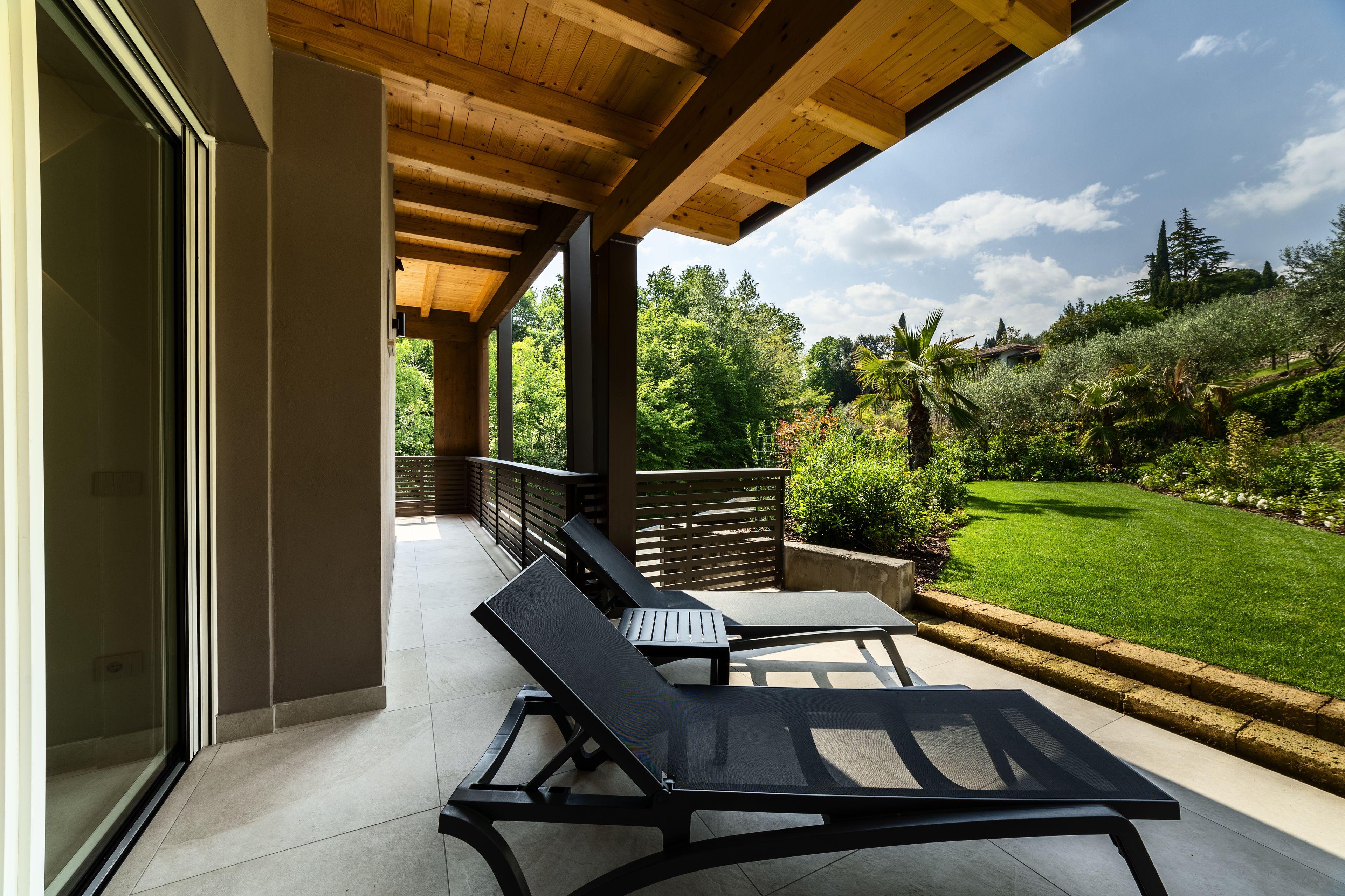 A covered patio with two lounge chairs, a wooden ceiling, and a view of a well-maintained garden.