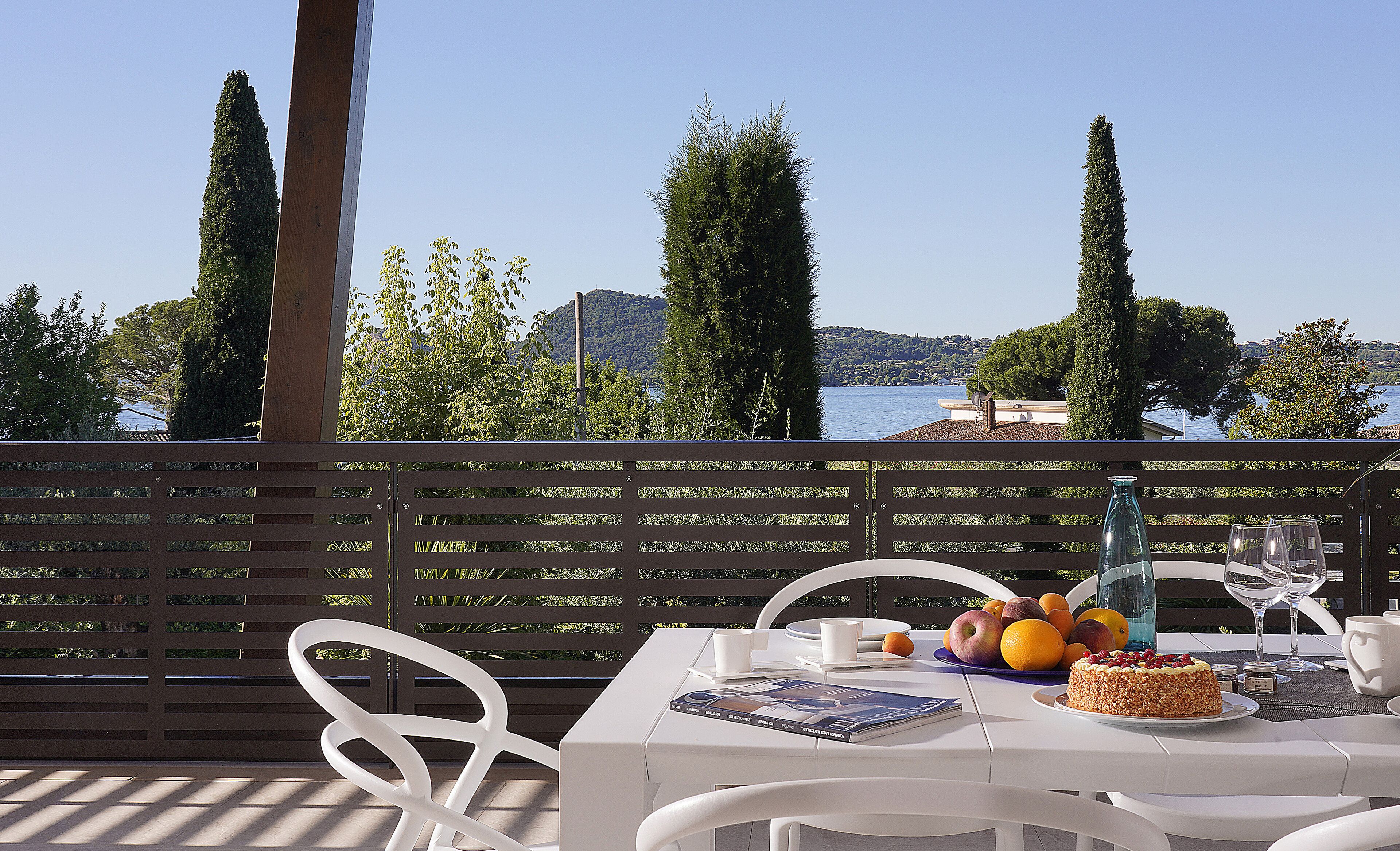 A table set with a fruit bowl, wine glasses, and a cake, overlooking a lake with trees and hills in the background.