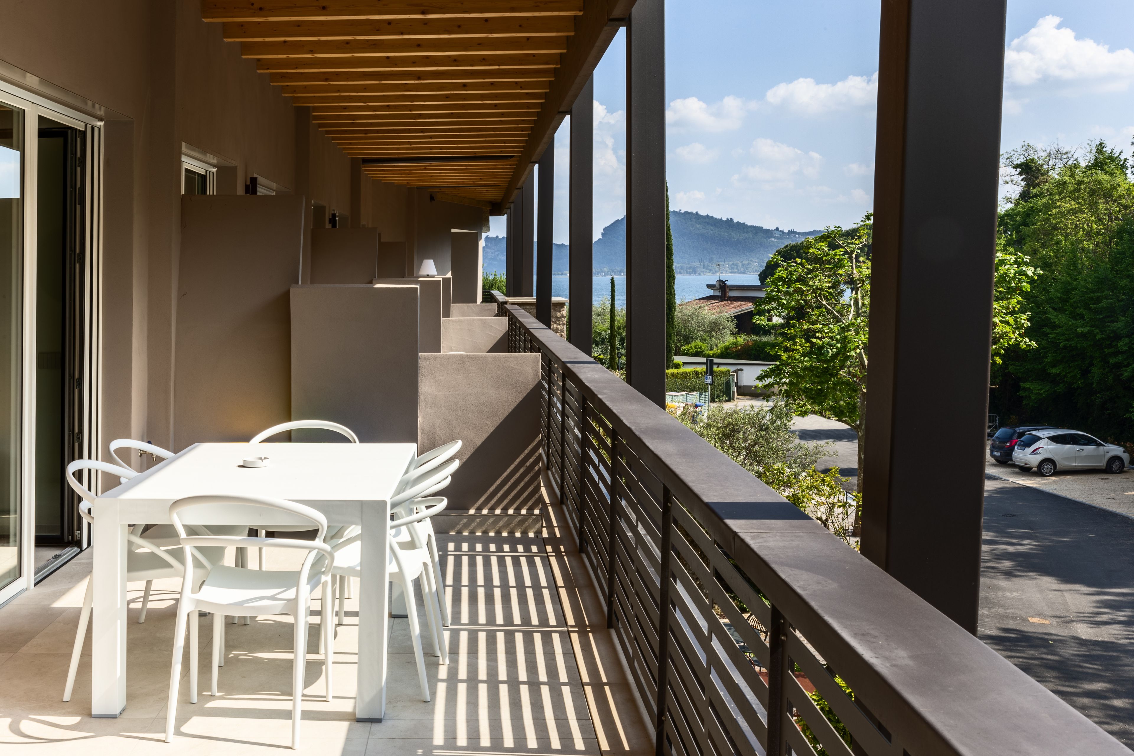 Un balcon avec une table et des chaises blanches, donnant sur un parking et un paysage montagneux.