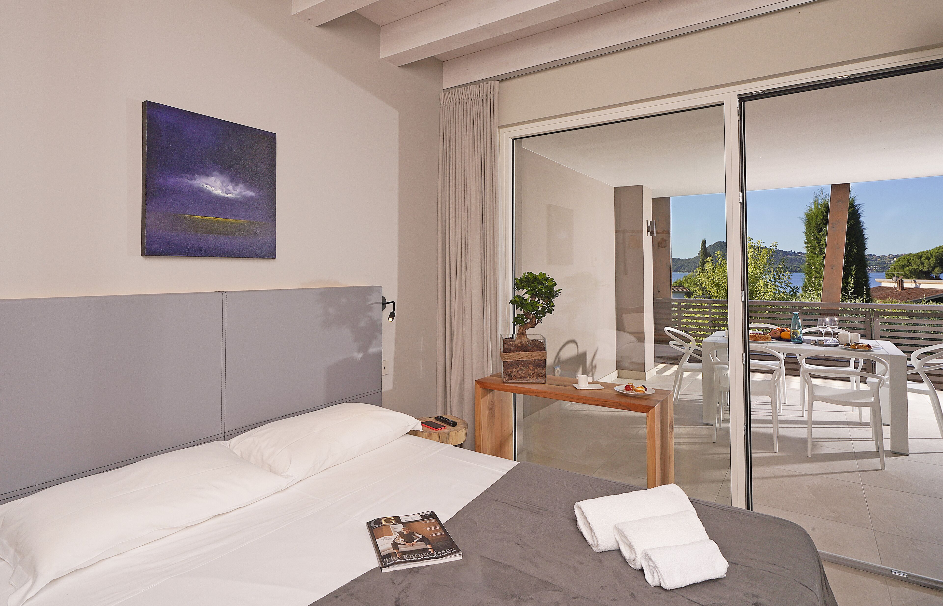 Une chambre d’hôtel moderne équipée d’un lit, d’une table de chevet, d’un magazine et offrant une vue sur la terrasse extérieure, avec une table et des chaises.