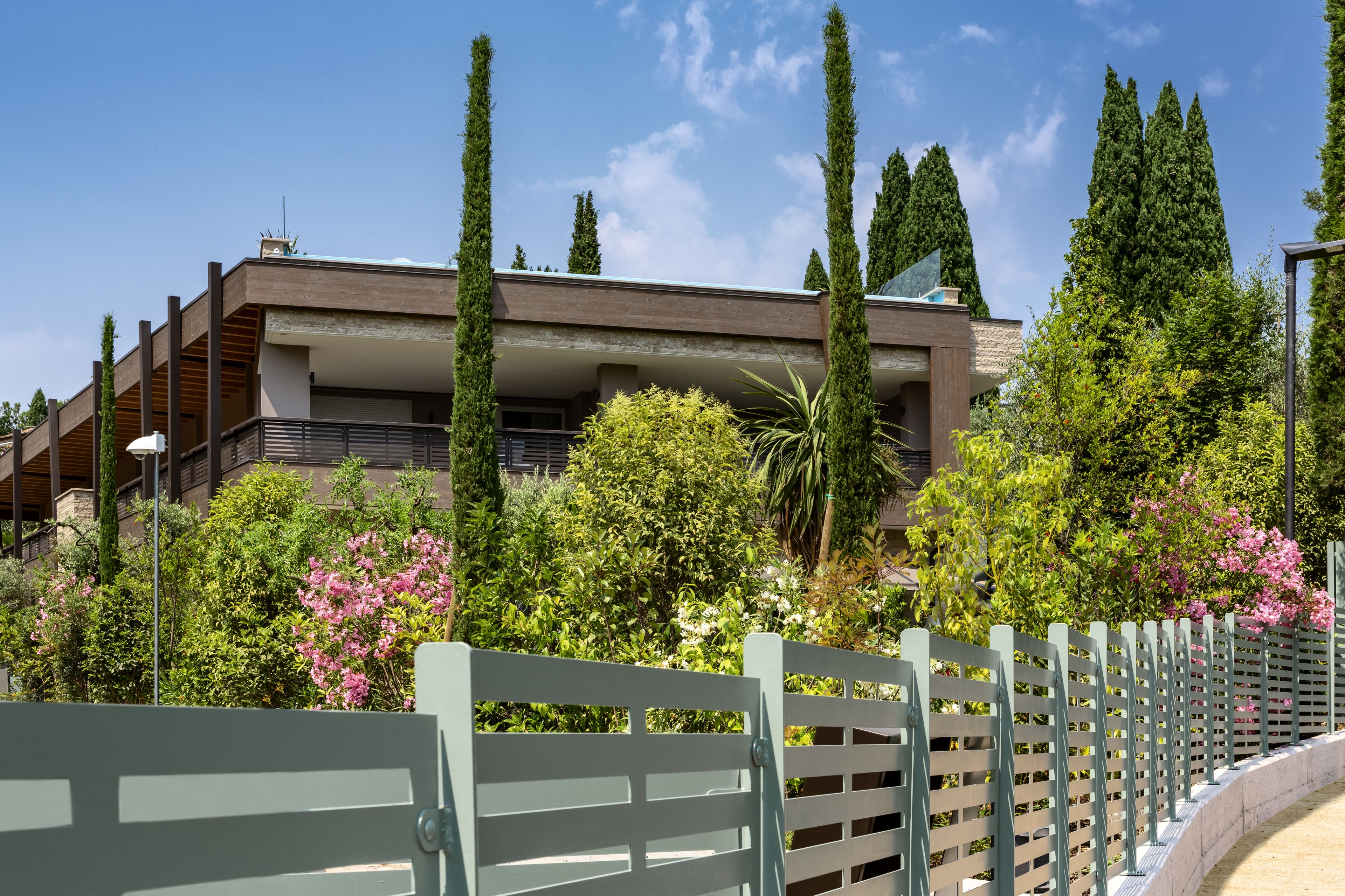 Un bâtiment moderne doté d’un toit plat, de nombreux balcons et d’un jardin environnant agrémenté de divers arbres et arbustes.