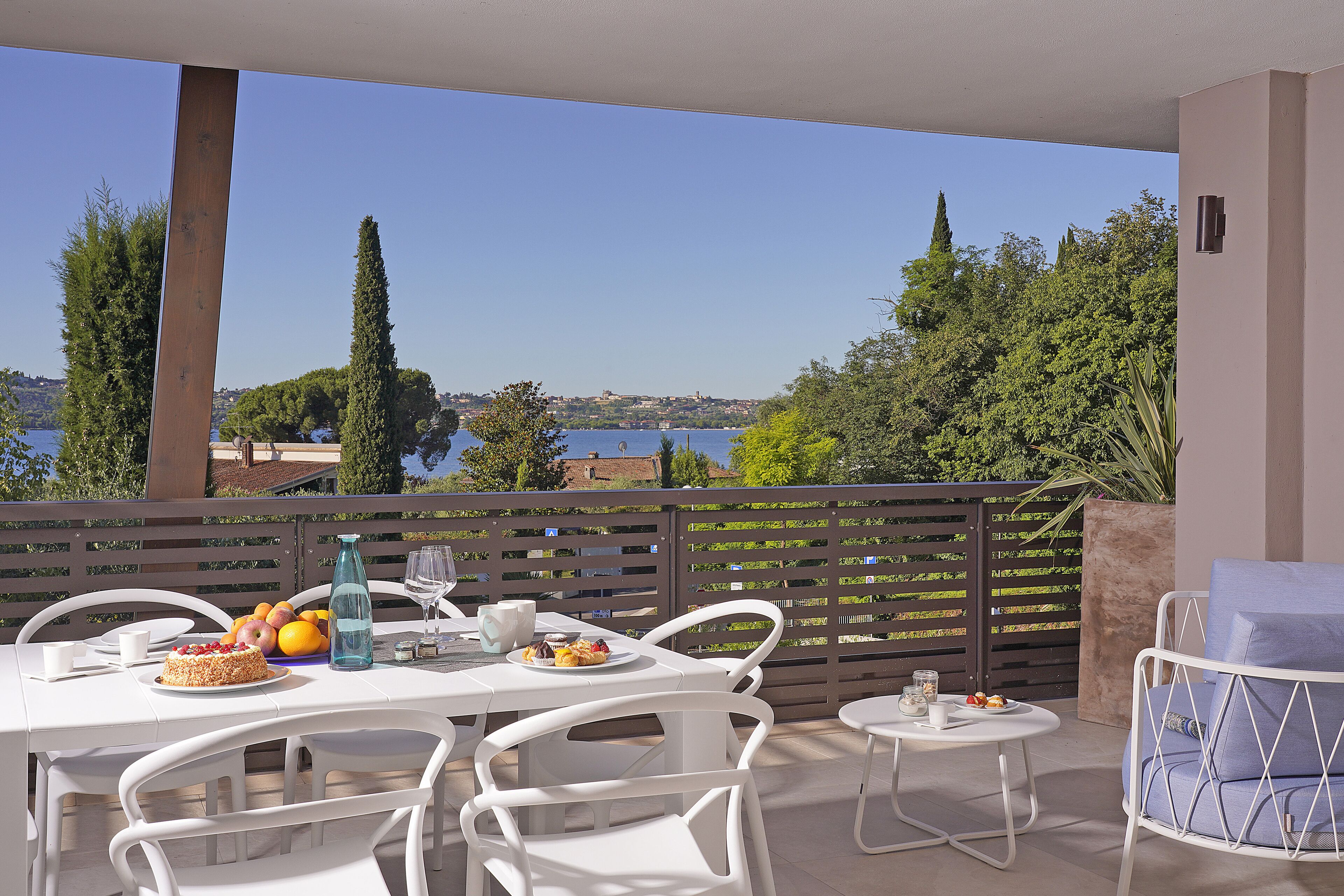 Una terraza con mobiliario blanco, una mesa preparada con comida y bebidas, y vistas al mar y a los árboles.