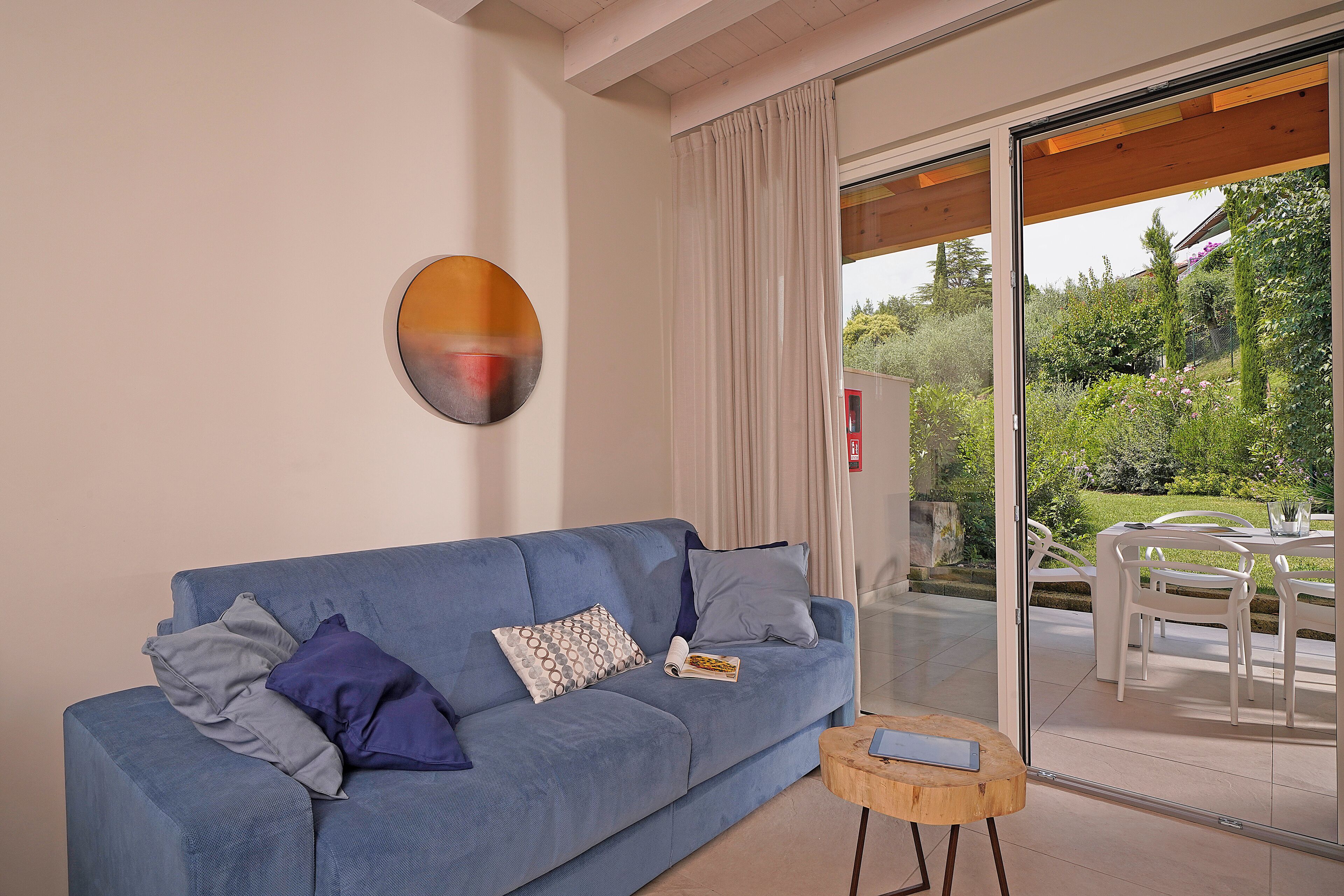 Un moderno salón con un sofá azul, una mesita de madera y un ventanal de vidrio que da a un patio exterior con mobiliario blanco.