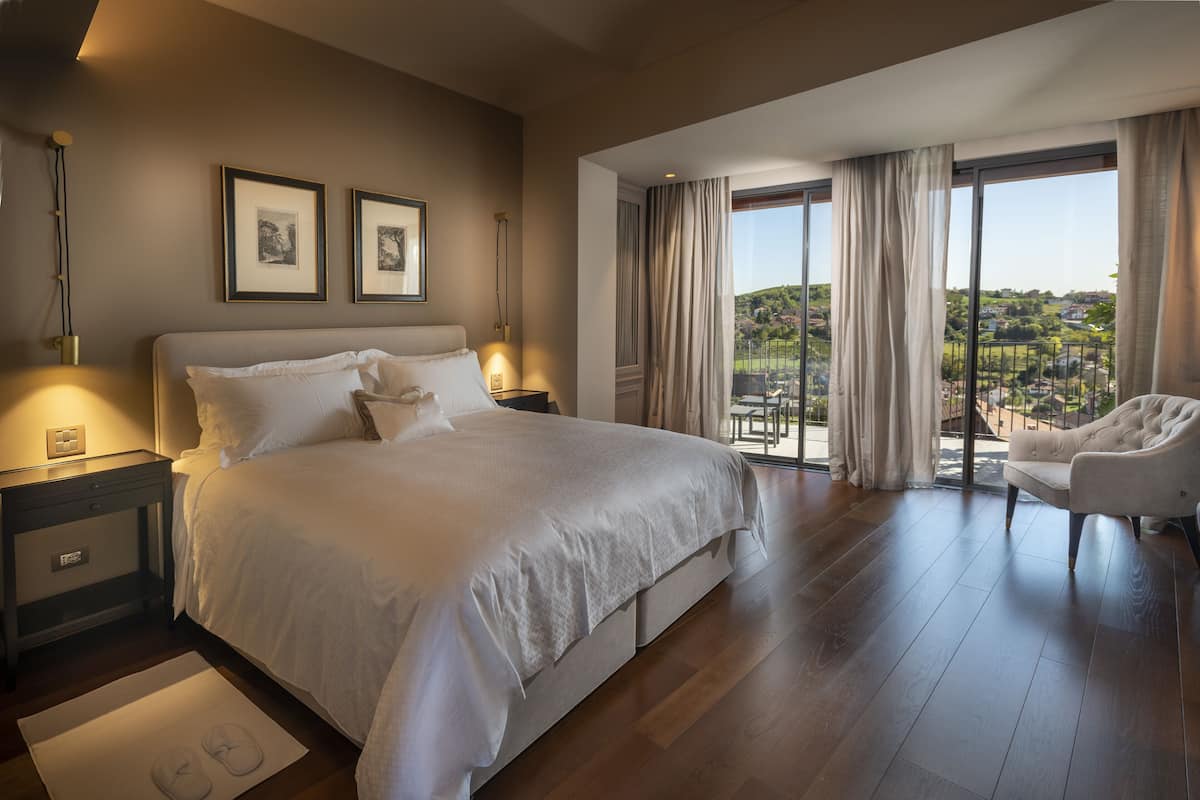 A spacious bedroom with a large bed, a nightstand, a chair, and a view of a balcony with a scenic landscape.