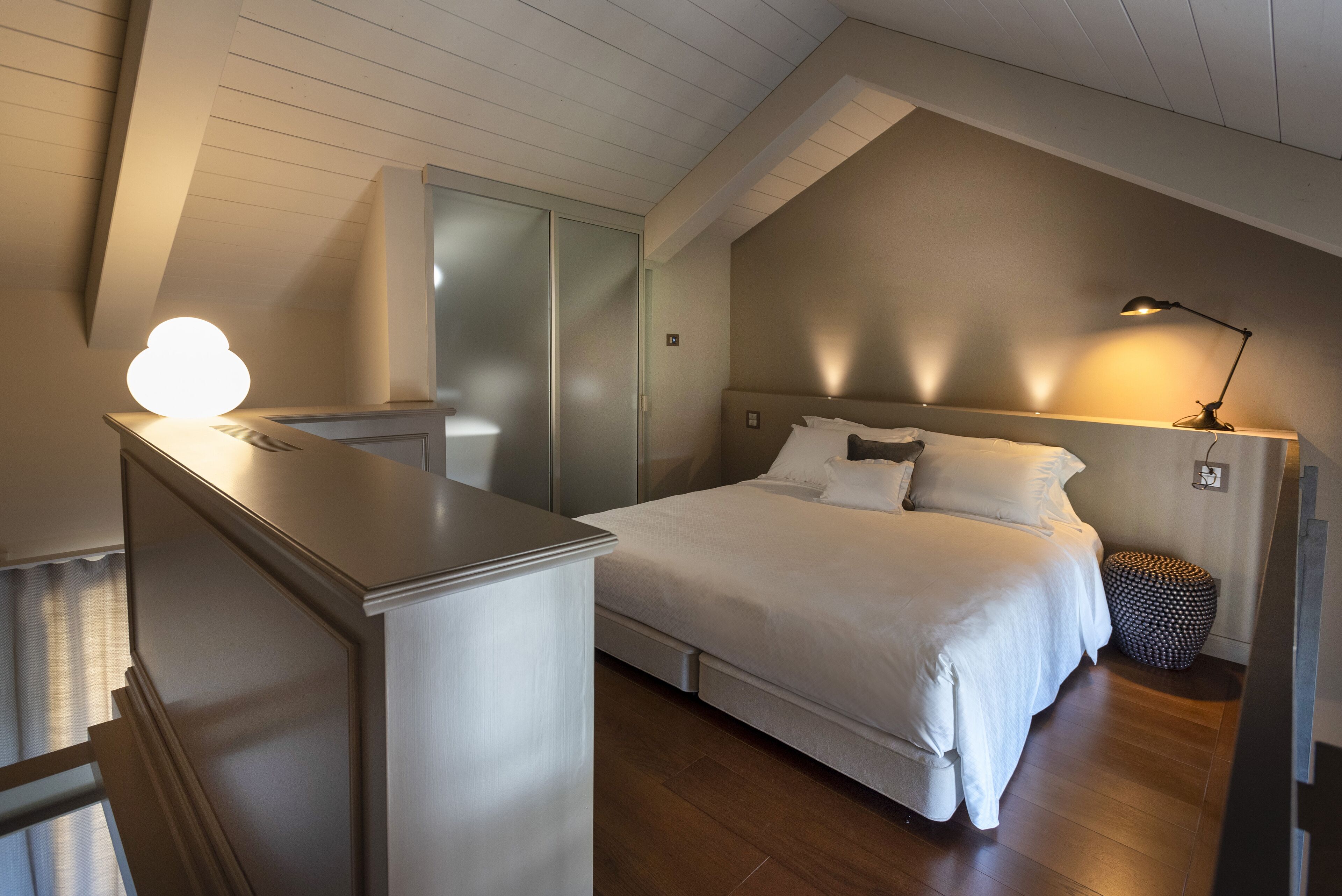 A bedroom with a bed, bedside table, lamp, and a small round ottoman.