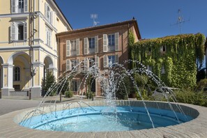 A public fountain with water jets in a circular pool, surrounded by buildings with classical architecture and a clear blue sky. - Dimora Cordero (Priocca)