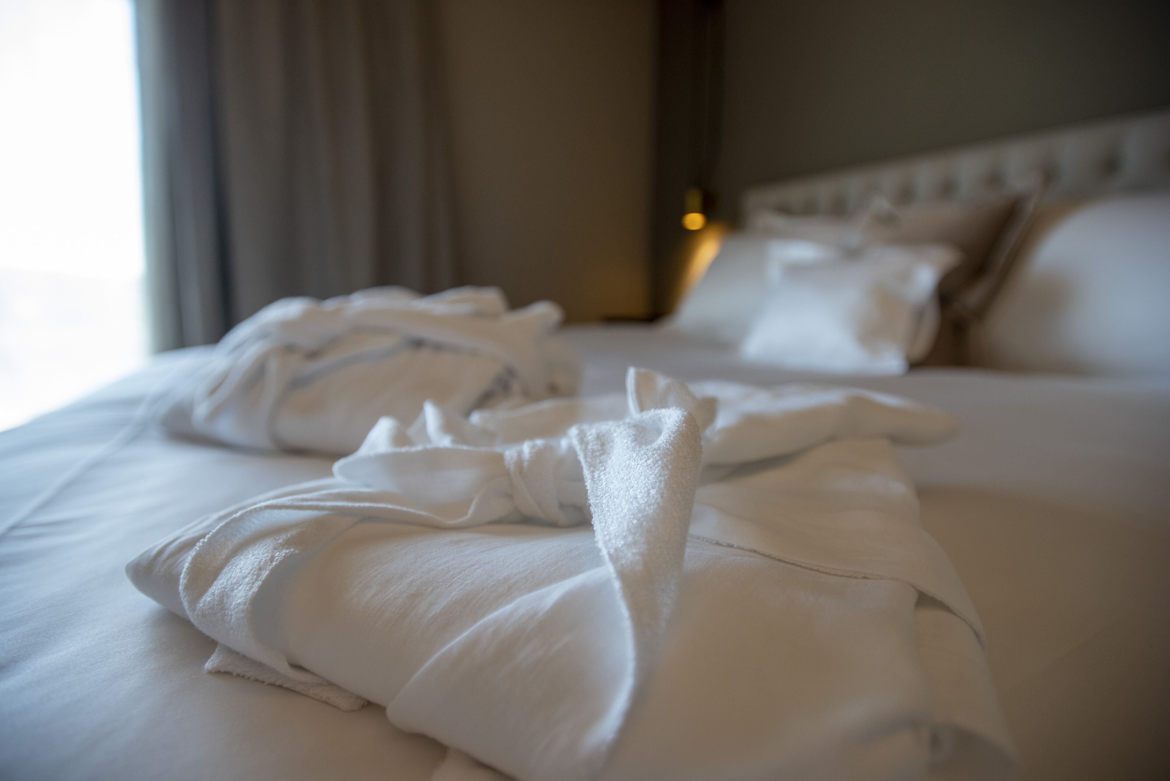 A neatly made bed with white linens and pillows, a headboard, and a window with curtains.