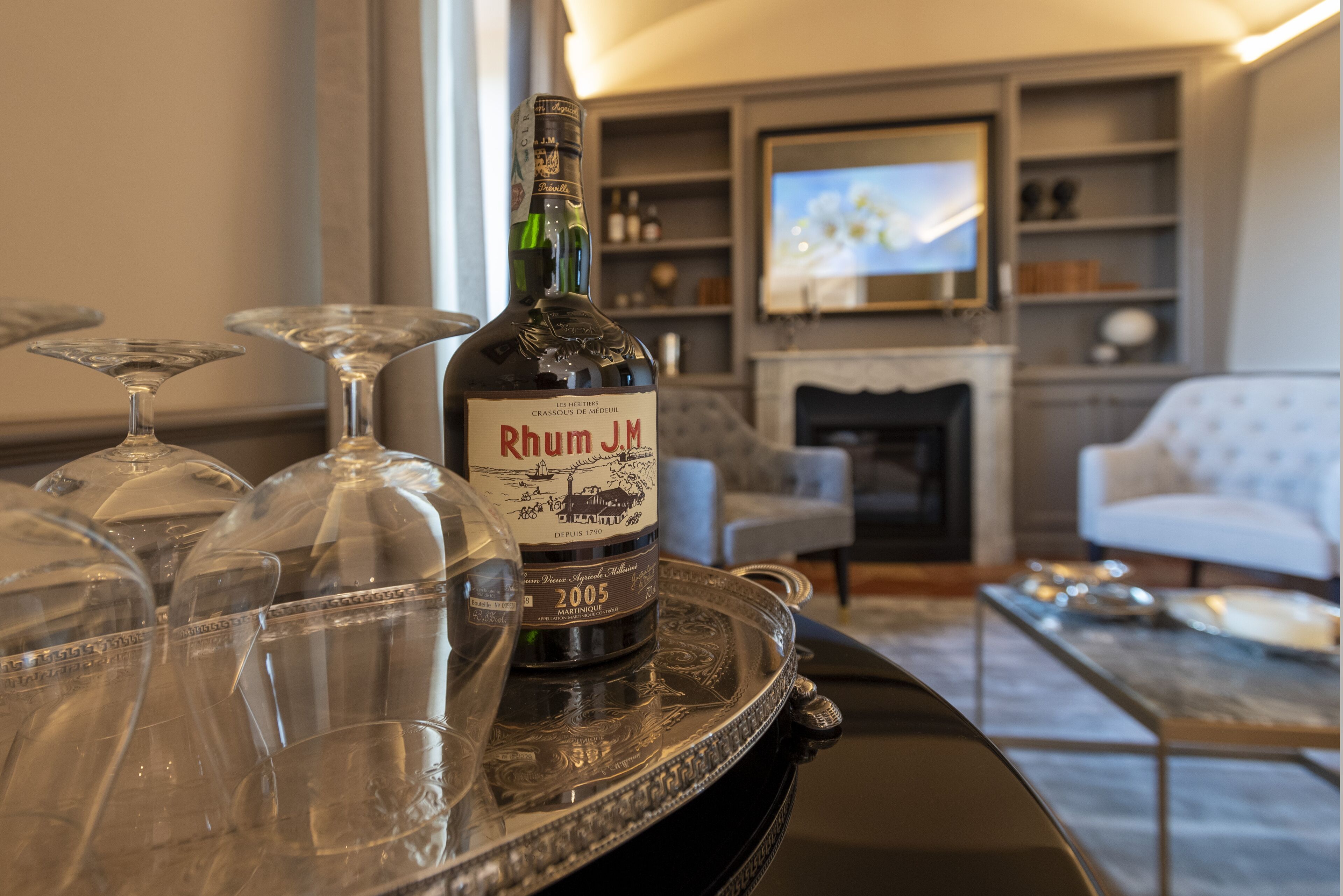 A bottle of Rhum J.M. 2005 on a tray with several empty glasses. The setting is a living room with a fireplace, a television, and a sofa.