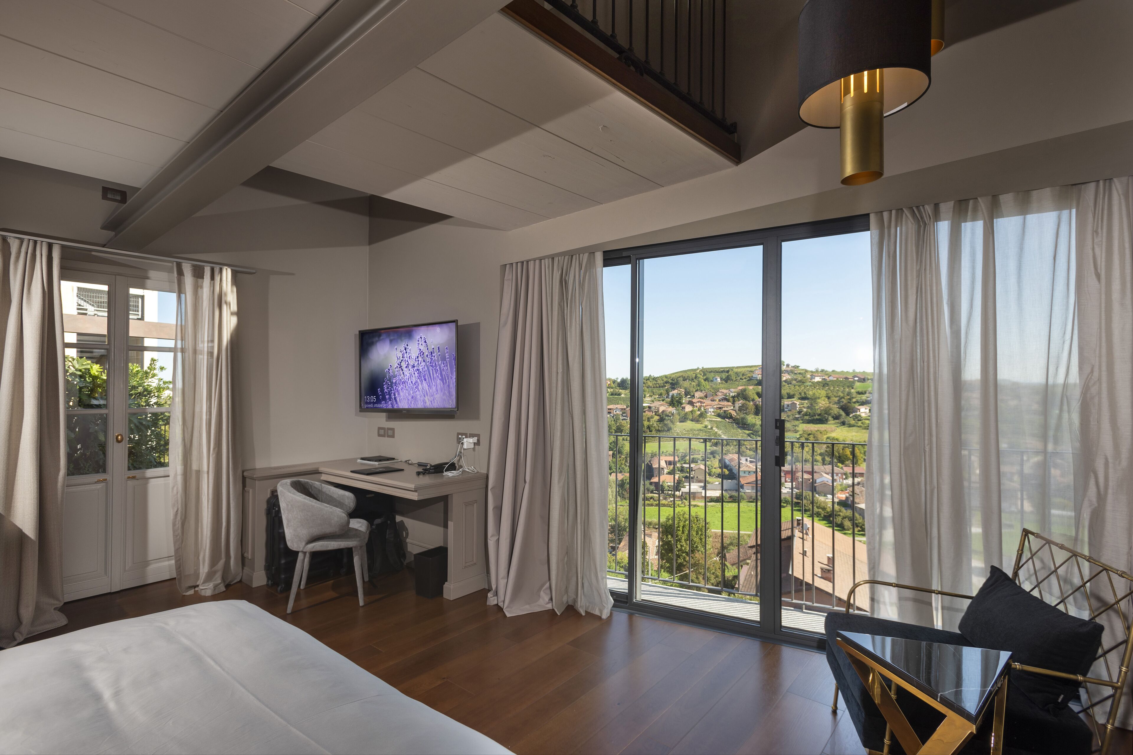 A modern hotel room with a large bed, a desk with a chair, a TV, and a balcony with a view of the countryside.