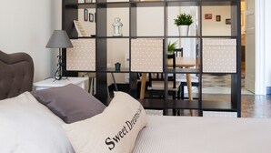 A bedroom with a bed, pillows, a nightstand, a lamp, a desk with a chair, and a shelving unit with plants and books.