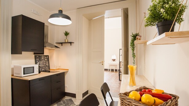 A modern kitchen with a microwave, black cabinets, and a hanging pendant light. There is a dining area with a table, chairs, and a basket of fruits.