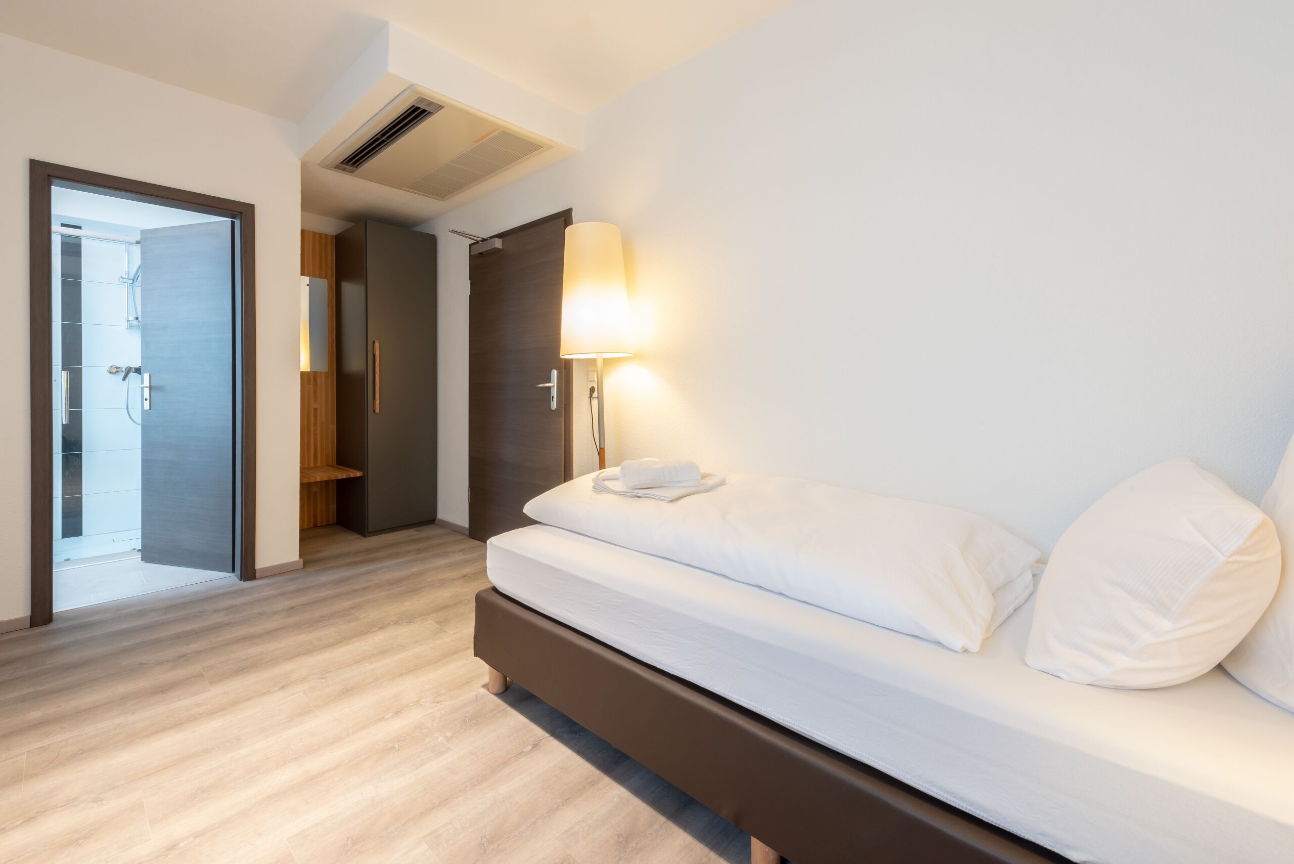 Habitació d'hotel moderna amb sòl de fusta, un llit amb roba blanca, una tauleta de nit amb una làmpada i una porta que dona a un bany.