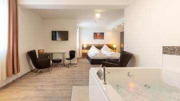 A hotel room with a large bathtub, a desk with a chair, a bed with pillows, and a flat-screen TV mounted on the wall.