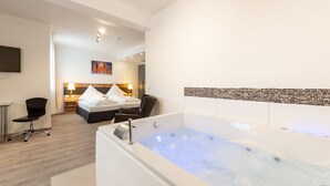 Una habitación de hotel moderna con una bañera grande, una cama con ropa blanca, un escritorio con una silla y un cuadro en la pared.