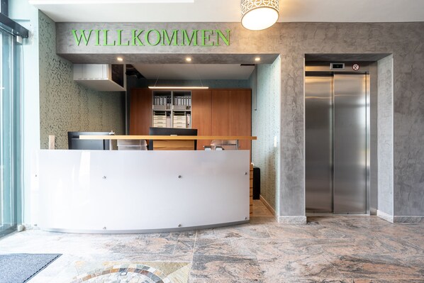 A reception area with a 'WILLKOMMEN' sign, a reception desk, a wooden cabinet, and an elevator. - Hotel in Laisen (Reutlingen)