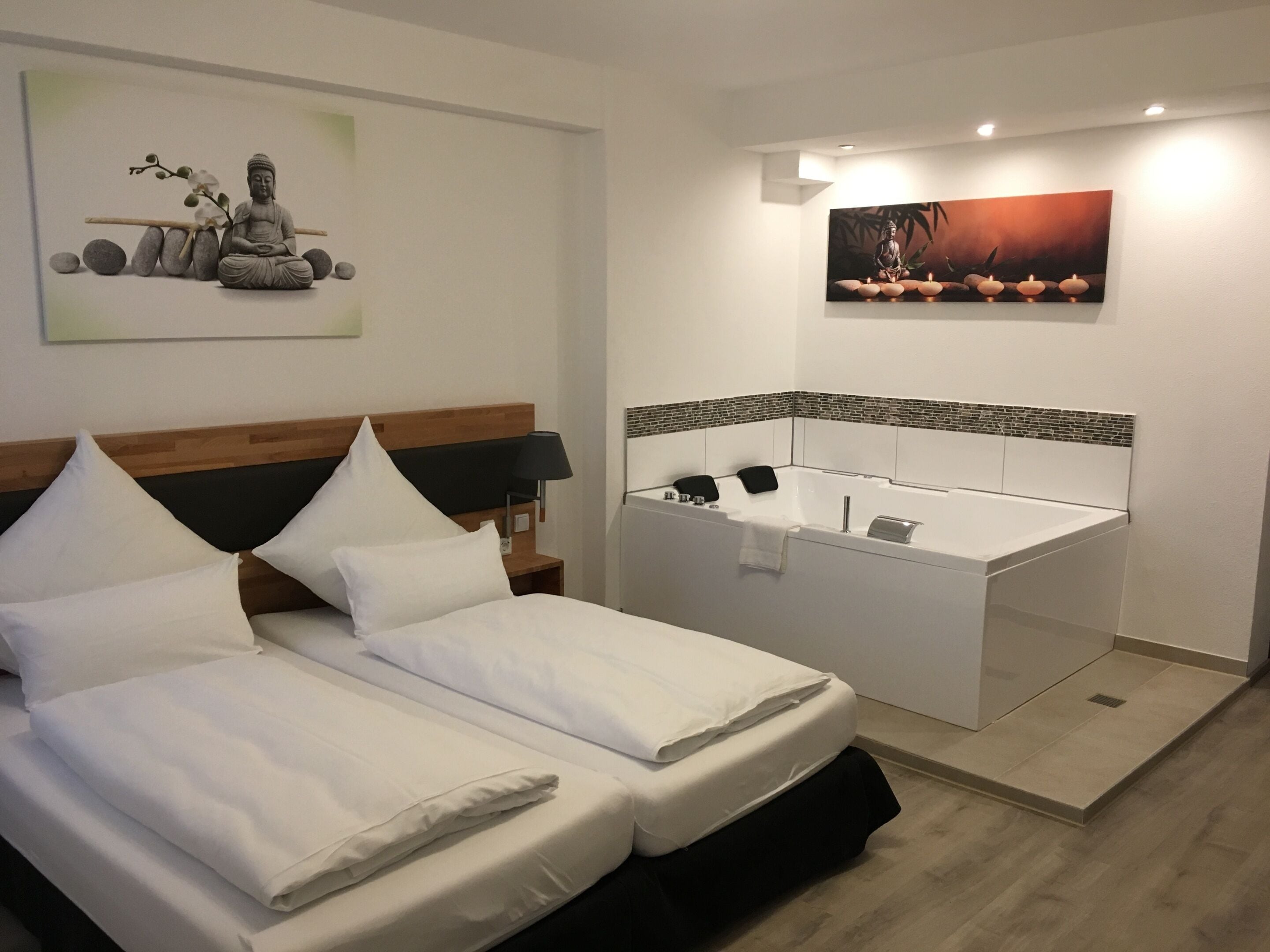 A hotel room with a bed, a nightstand, a wall-mounted lamp, a painting, and a bathtub with a towel.