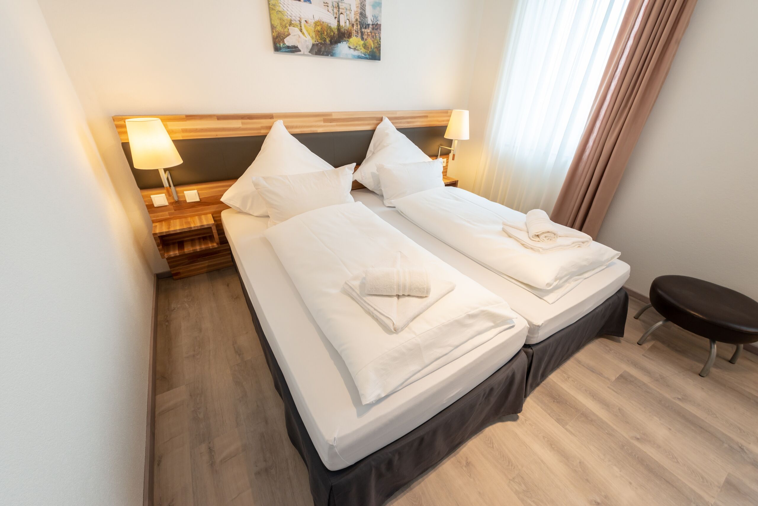 Une chambre d’hôtel avec deux lits, des têtes de lit en bois, du linge de lit blanc, une table de chevet avec une lampe et un tableau au mur.