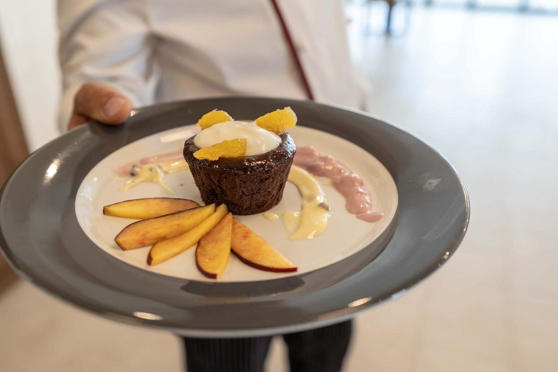 Eine Person hält einen Teller mit Dessert, darunter ein Cupcake mit Schlagsahne und Früchten sowie Apfelscheiben mit Sauce.