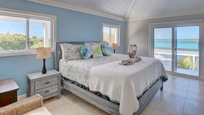 A bedroom with a bed, nightstands, a lamp, and large windows offering a view of the sea.