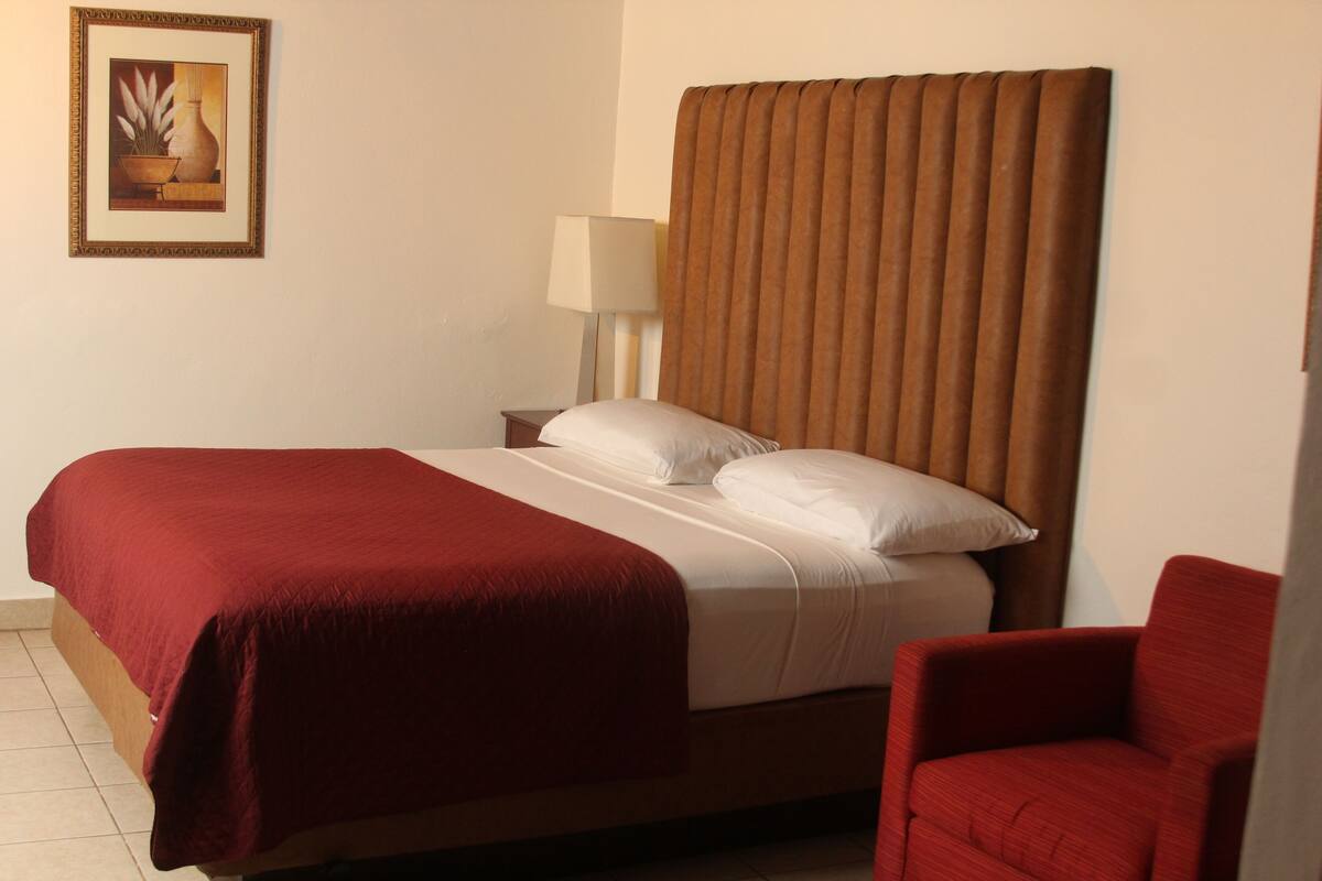 A hotel room with a large bed, a red chair, a nightstand, a lamp, and a framed picture on the wall.