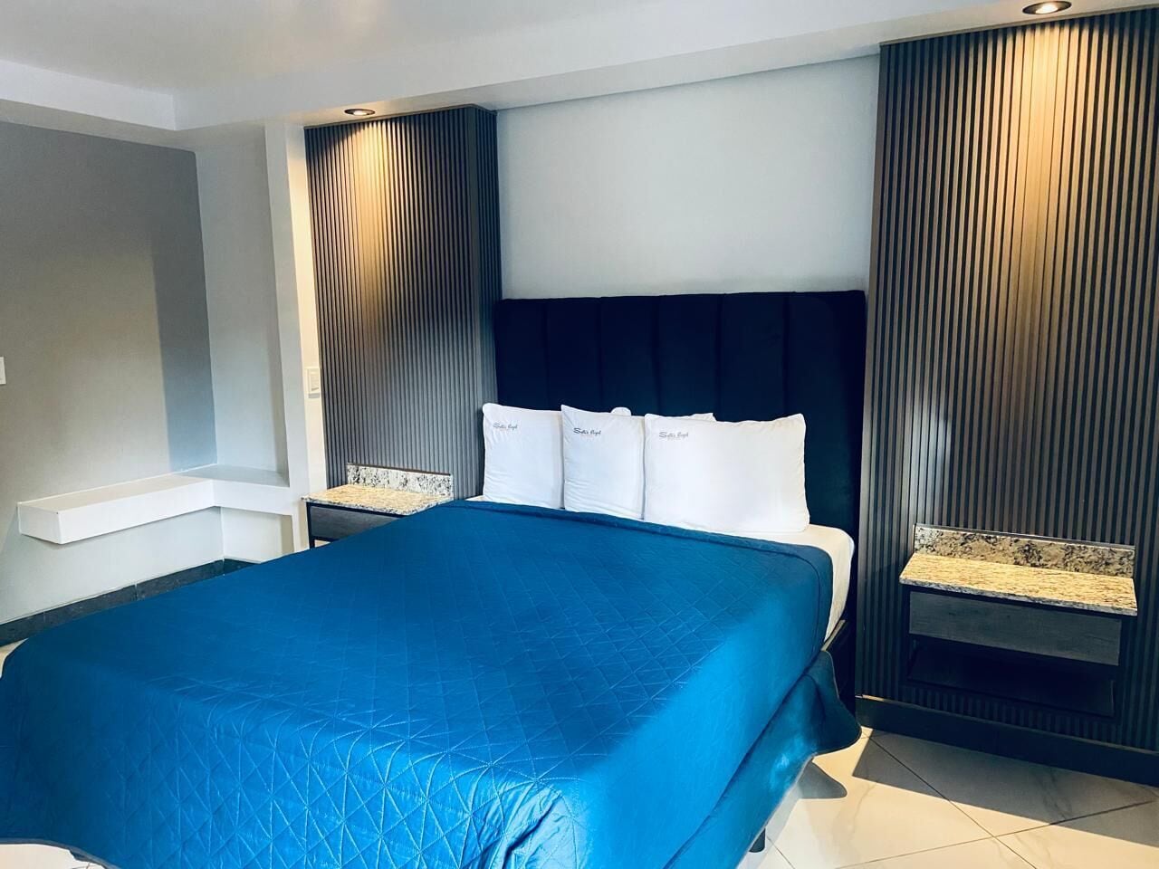 Una habitación de hotel moderna con una cama grande, un cabecero con listones verticales y una mesita de noche pequeña con una lámpara.