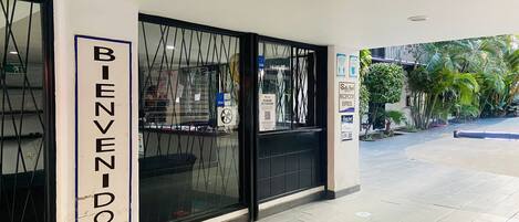 A hotel entrance with a 'BIENVENIDOS' sign, a reception desk, and a glass door.