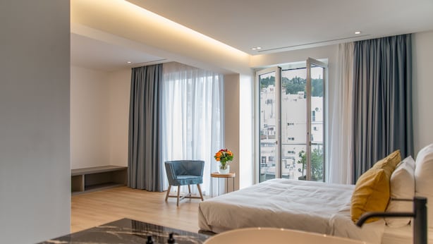 Et moderne hotelværelse med en stor seng, et siddeområde med en stol, et lille bord med en vase blomster og udsigt til bygninger gennem store vinduer.