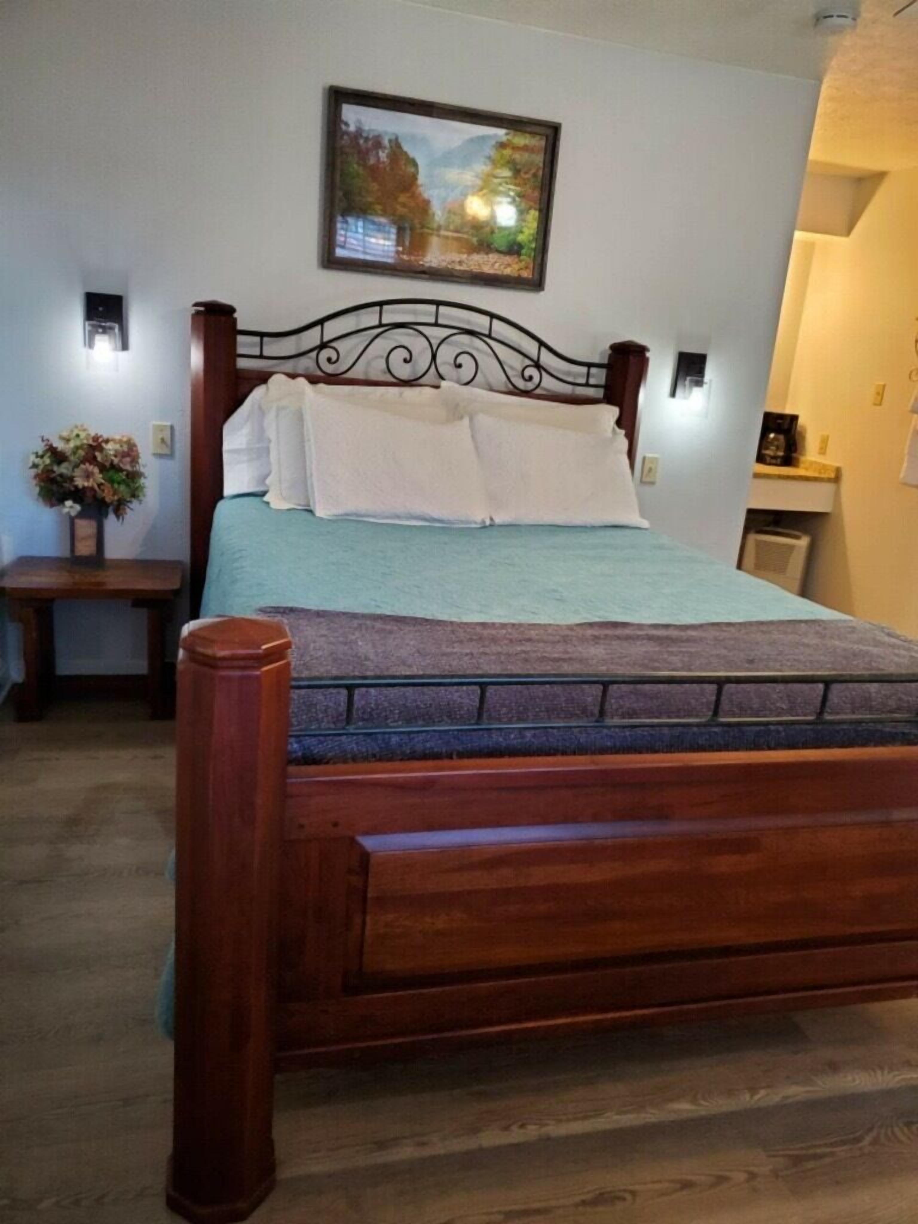 A bedroom with a wooden bed frame, a blue mattress, and a white pillow. There is a small bedside table with a vase of flowers. A framed picture hangs on the wall above the bed.