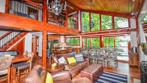 A spacious living area with a high ceiling, wooden beams, a large chandelier, and a view of the outdoors through large windows.
