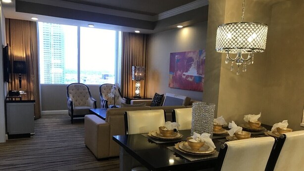 Une salle à manger d’hôtel avec une table noire dressée pour six personnes, entourée de chaises de couleur beige. Un lustre est suspendu au-dessus de la table, et une grande fenêtre avec des rideaux est visible à l’arrière-plan.