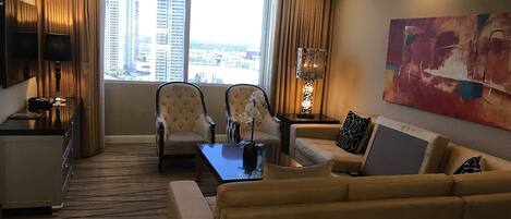 A hotel room with a large window, two armchairs, a sofa, a coffee table, a TV, and a painting on the wall.