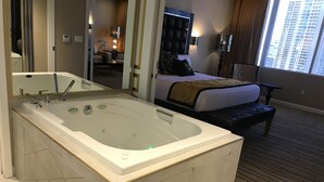 Une chambre d’hôtel dotée d’une grande baignoire, d’un lit avec une tête de lit, d’une table de chevet et offrant une vue sur le paysage urbain par la fenêtre.