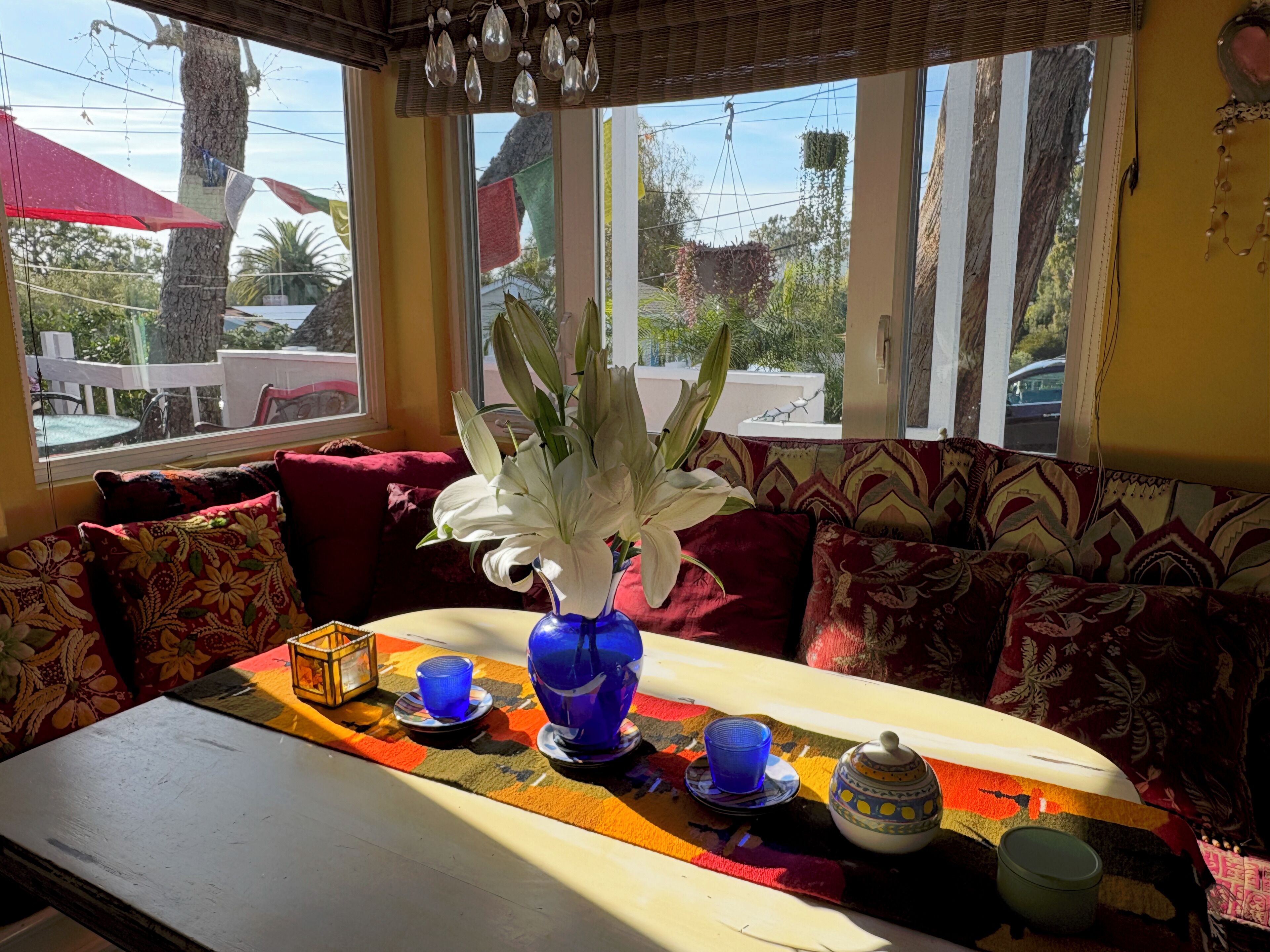 A cozy seating area with a table set for tea, a vase with white flowers, and a view of the outside through large windows.