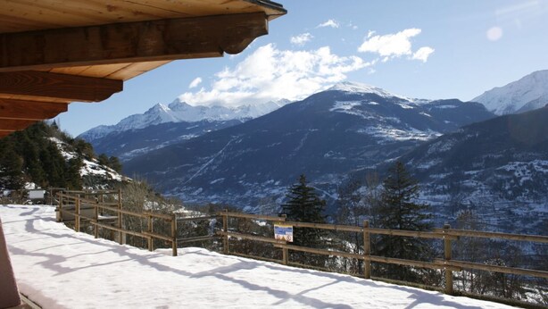 Un balcon en bois donnant sur un paysage montagneux enneigé, sous un ciel dégagé.