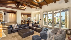 A spacious living room with a large sectional sofa, ottomans, and a fireplace. There is a kitchen in the background and a patio with outdoor furniture. - Soquel Villa - 180 views of Monterey Bay w/ infinity pool, spa & movie theater (Soquel)