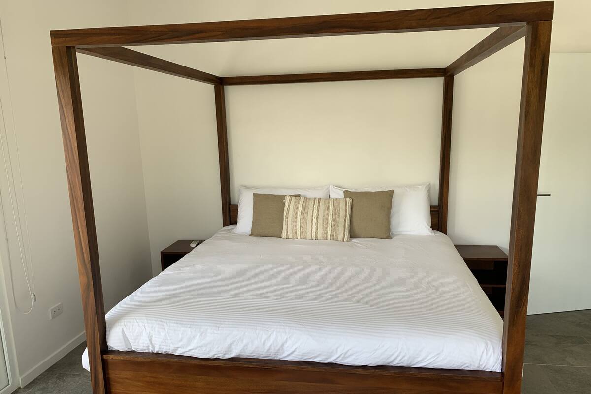 A four-poster bed with white bedding and two pillows, a wooden nightstand, and a plain wall.