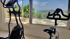 A home gym with two exercise bikes and an elliptical machine, large windows with a view of greenery and a cloudy sky. - Aves Hotel & Resort - Retreat Center (Cóbano)