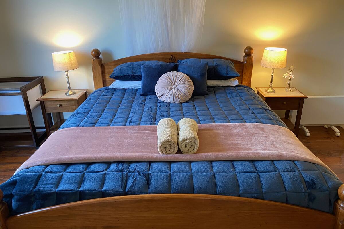 Una cama bien tendida con colcha azul, dos almohadas y una manta rosa. Hay dos lámparas de noche sobre mesitas de madera.