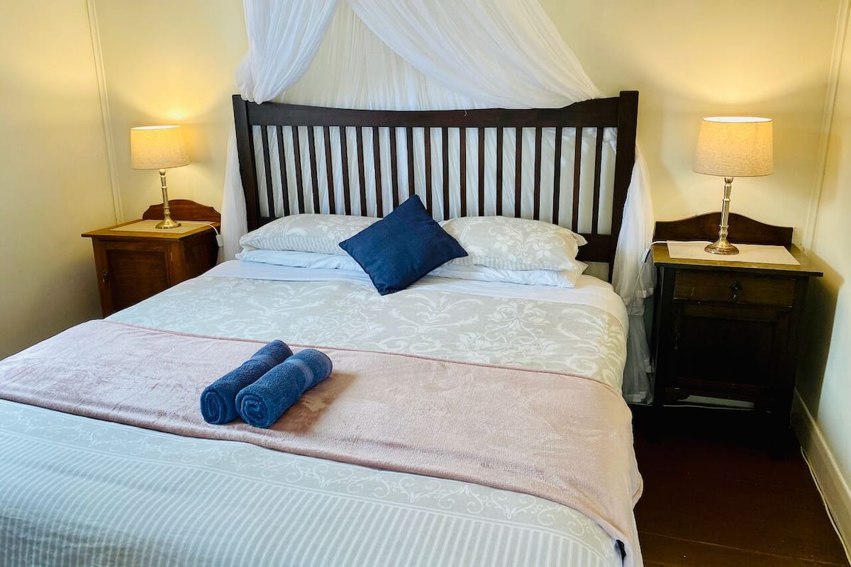 Una cama bien hecha con dosel, dos mesitas de noche con lámparas y un par de toallas enrolladas sobre la cama.