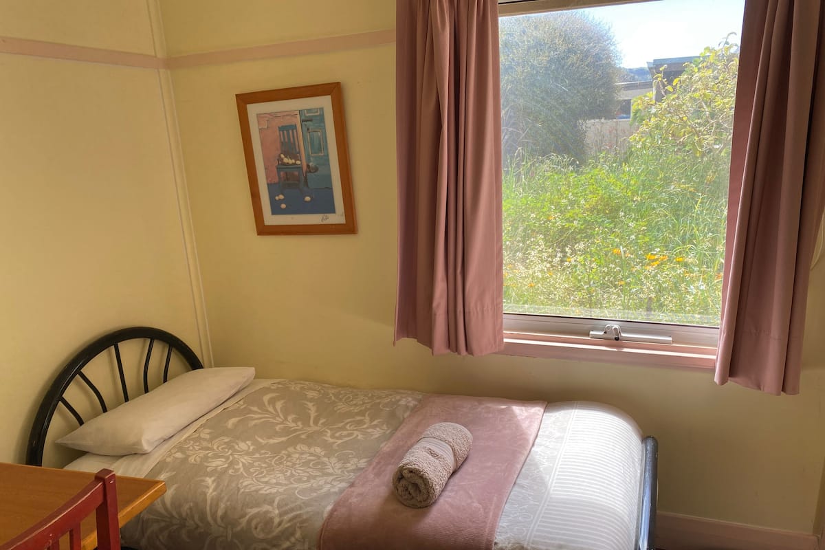 Una cama individual con una toalla enrollada, una silla de madera, un cuadro en la pared y una ventana con cortinas.