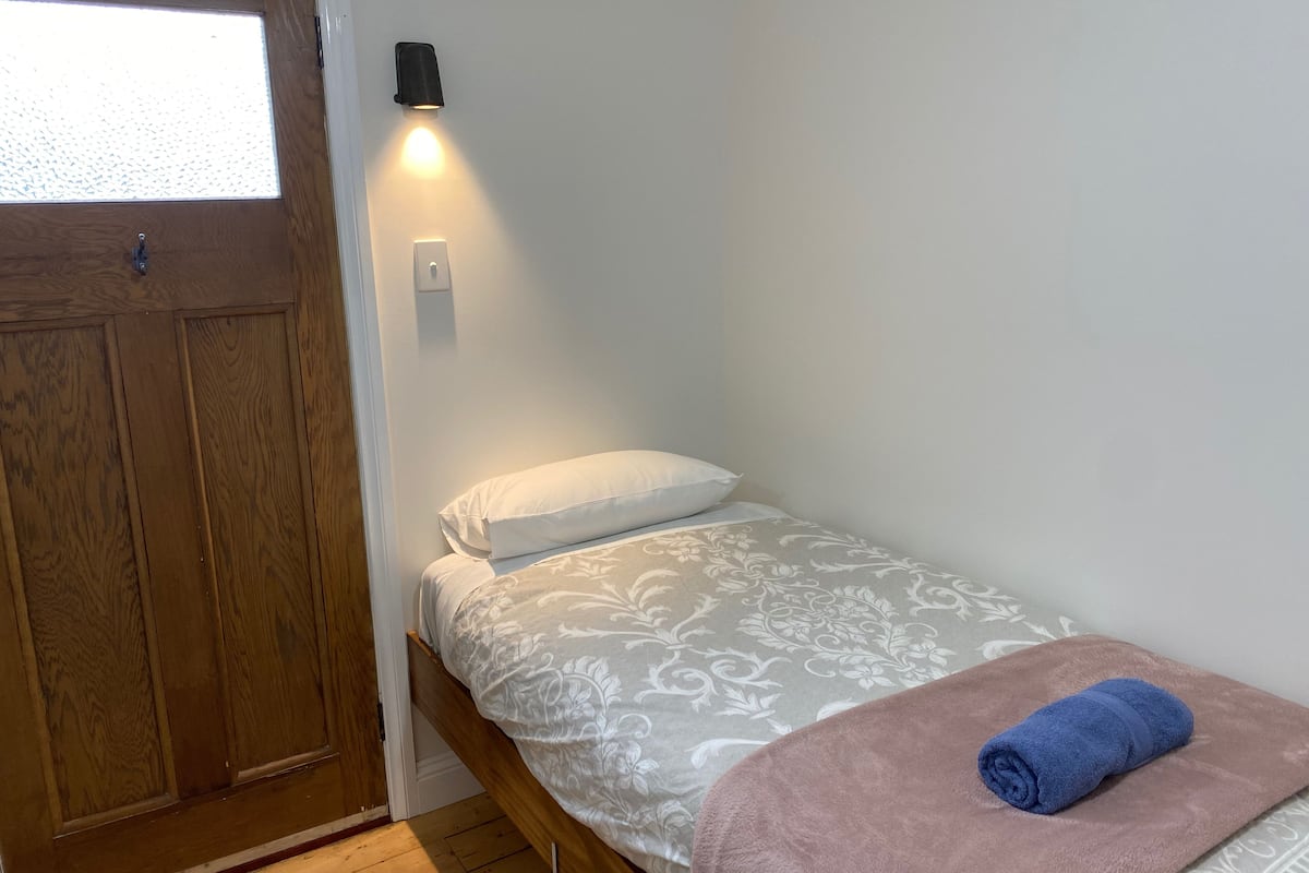 Habitación pequeña con cama individual, armario de madera, luz de pared y una toalla azul sobre la cama.