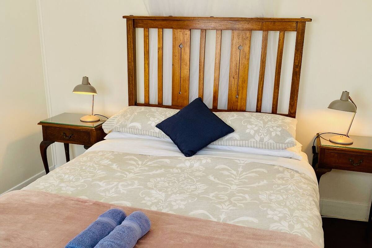 Una cama bien hecha con cabecera de madera, dos mesitas de noche con lámparas y una colcha rosa con toallas enrolladas.